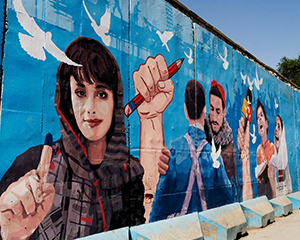 Wandgemälde mit einer Frau, einer Hand, die einen Bleistift hochhält, sich umarmenden Männern, einem Mikrofon, einer Frau und einem Kind sowie Friedenstauben.