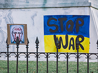 Image shows fence with a sign of Putin with red paint on his face and a sign of a Ukrainian flag that reads "Stop War"